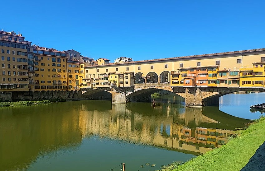 Ponte Vecchio, the medieval stone bridge in Florence, with shops along both sides reflected in the Arno River on a clear day.