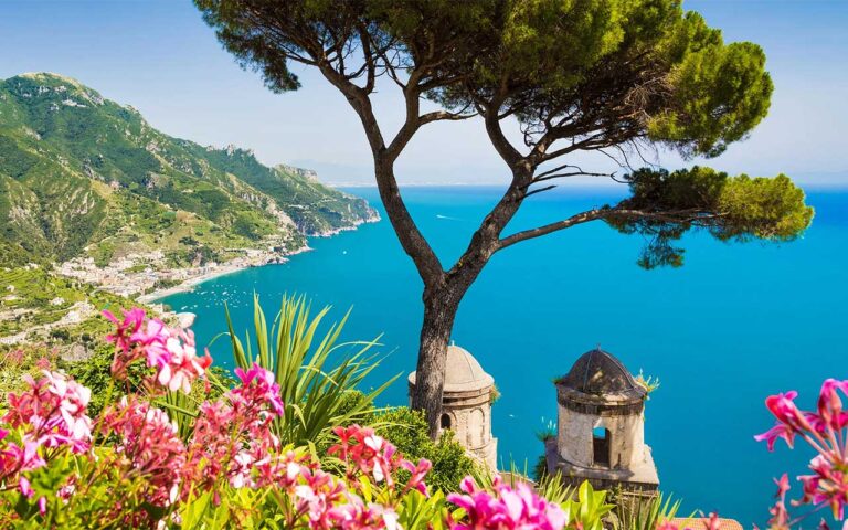 Panoramic view of the Amalfi Coast from Villa Rufolo in Ravello, featuring a lone pine tree, colorful flowers, historic domes, and the turquoise Mediterranean Sea.