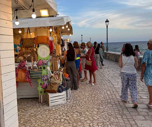 People shopping at a Sperlonga night market stall selling handmade bags, hats, and local crafts by the seaside.